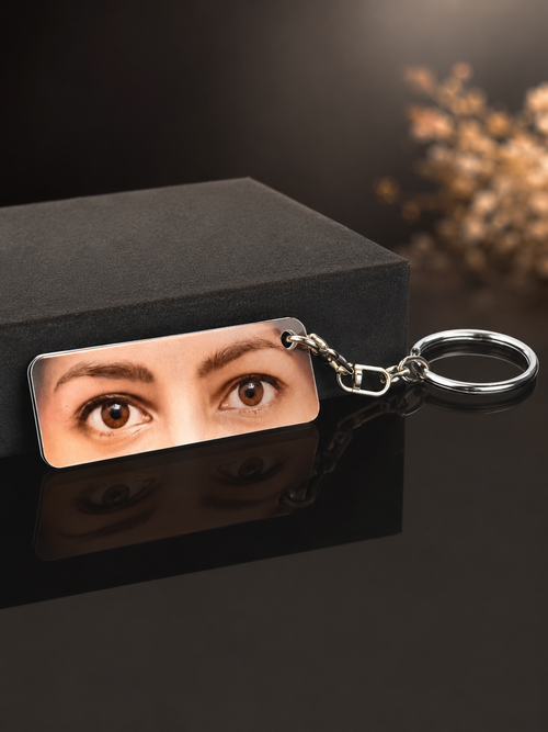 A rectangular metal keychain with a custom printed photo of a woman's eyes, attached to a silver split ring, displayed on a black box against a dark background.