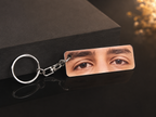 A rectangular acrylic keychain featuring a close-up photo of a person's eyes, attached to a silver metal split ring and clasp, displayed on a dark reflective surface next to a black box.
