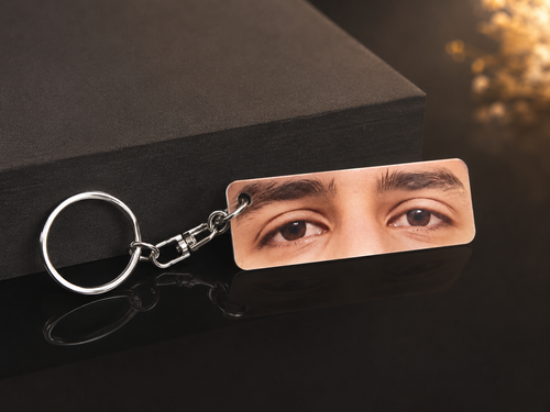 A rectangular acrylic keychain featuring a close-up photo of a person's eyes, attached to a silver metal split ring and clasp, displayed on a dark reflective surface next to a black box.