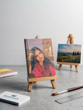 Small wooden easel with a portrait painting on a gray surface, with another painting and art supplies in the background, made by Kustomade, India.
