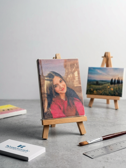 Small wooden easel with a portrait painting on a gray surface, with another painting and art supplies in the background, made by Kustomade, India.